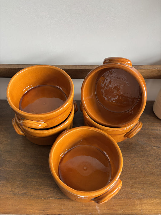 Five small glazed ceramic bowls with side handles, stacked and arranged on a wooden surface against a light wall.