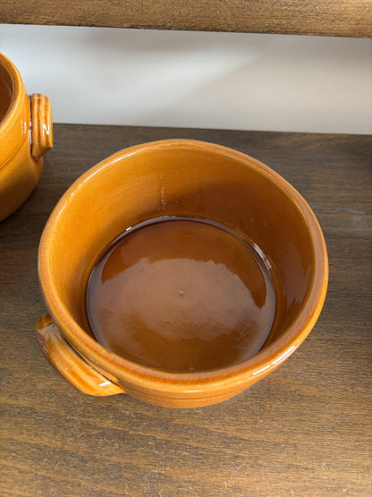 A close-up of a small brown ceramic bowl with a glossy amber-orange glazed finish and a side handle, sitting on a dark wooden surface.