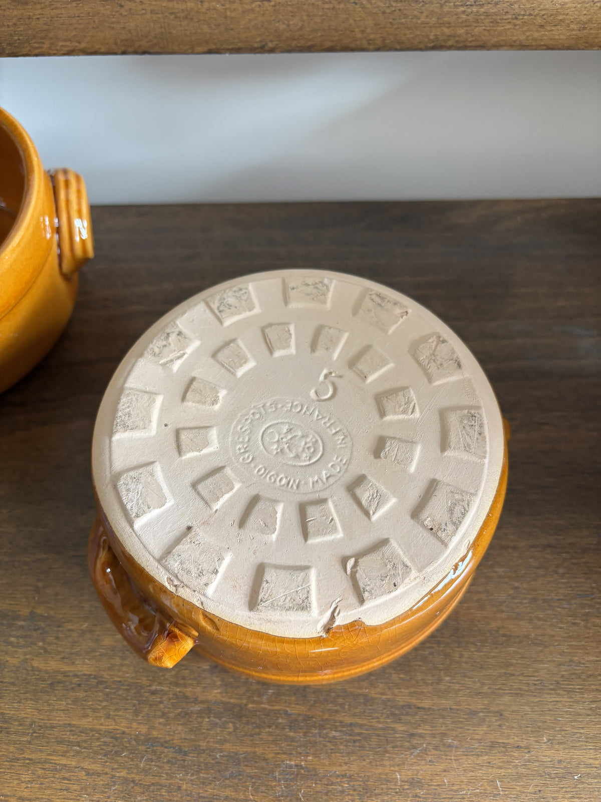 A close-up of the bottom of a small round ceramic pot showing a cream-colored foot ring and a glossy amber-orange glazed body, placed on a wooden surface.