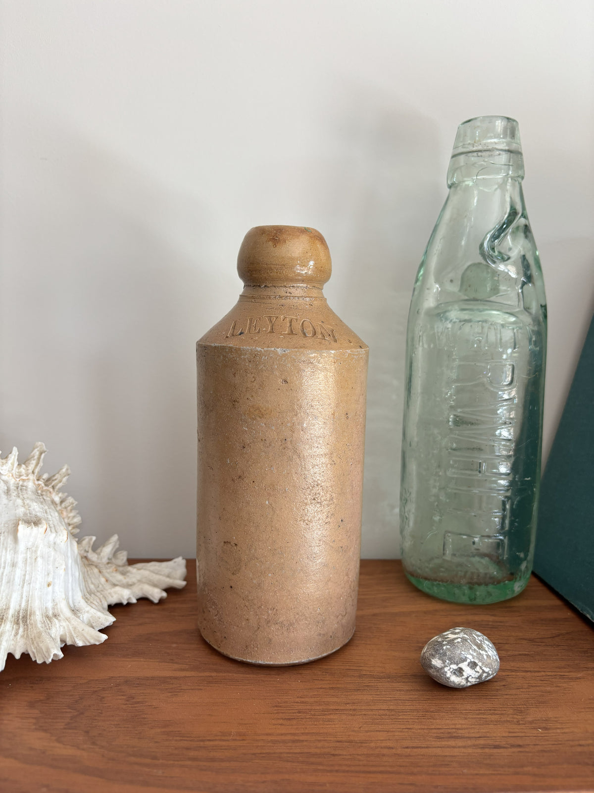 A vintage brown stoneware bottle with a rounded shoulder and embossed brand text, standing on a wooden surface next to a clear glass bottle and a white seashell.
