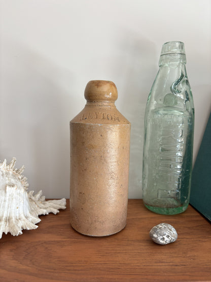 A vintage brown stoneware bottle with a rounded shoulder and embossed brand text, standing on a wooden surface next to a clear glass bottle and a white seashell.