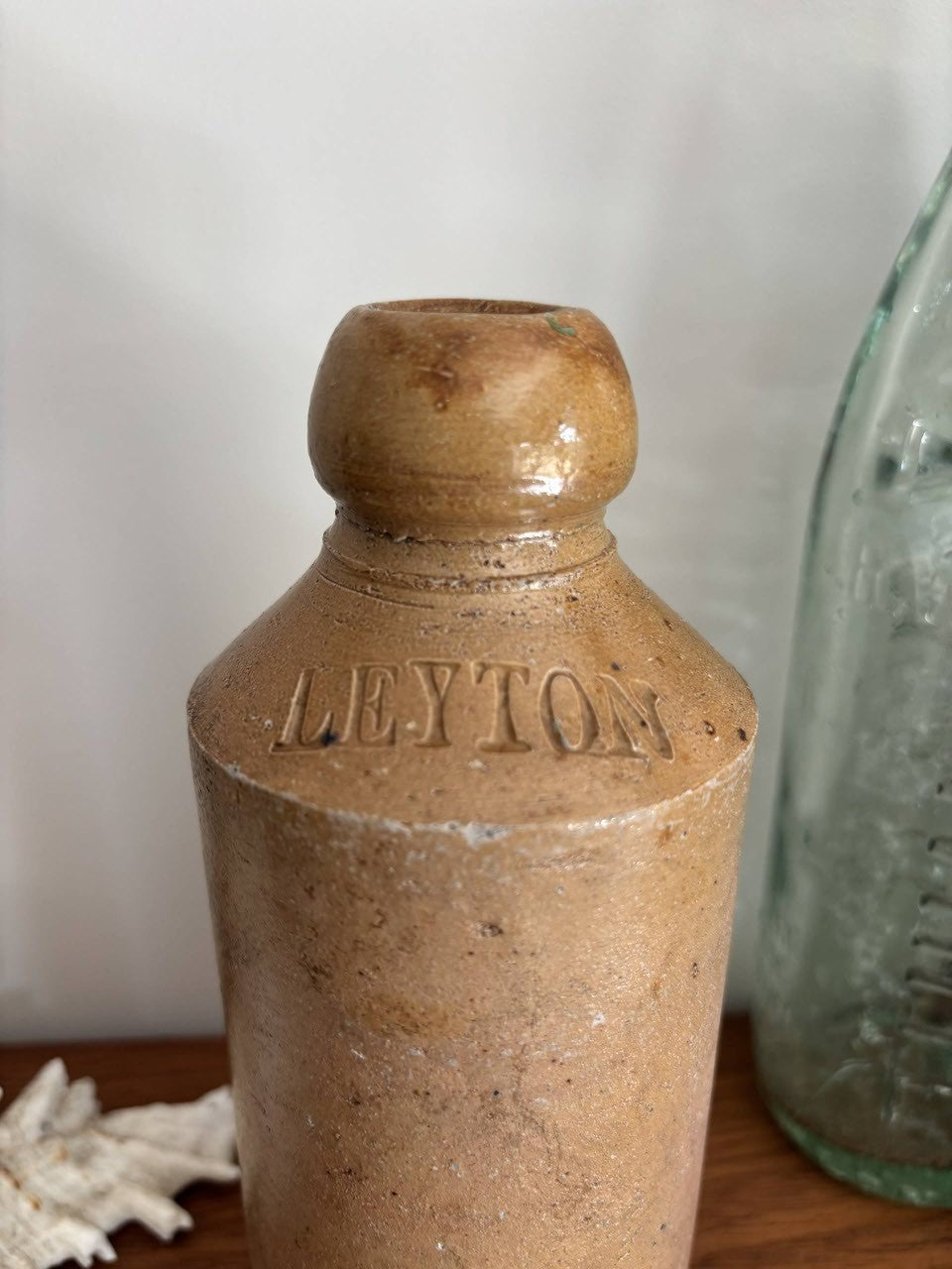 A vintage brown stoneware bottle with 'LEYTON' embossed on the front, displayed on a wooden surface next to a white seashell and a blurred green glass bottle.