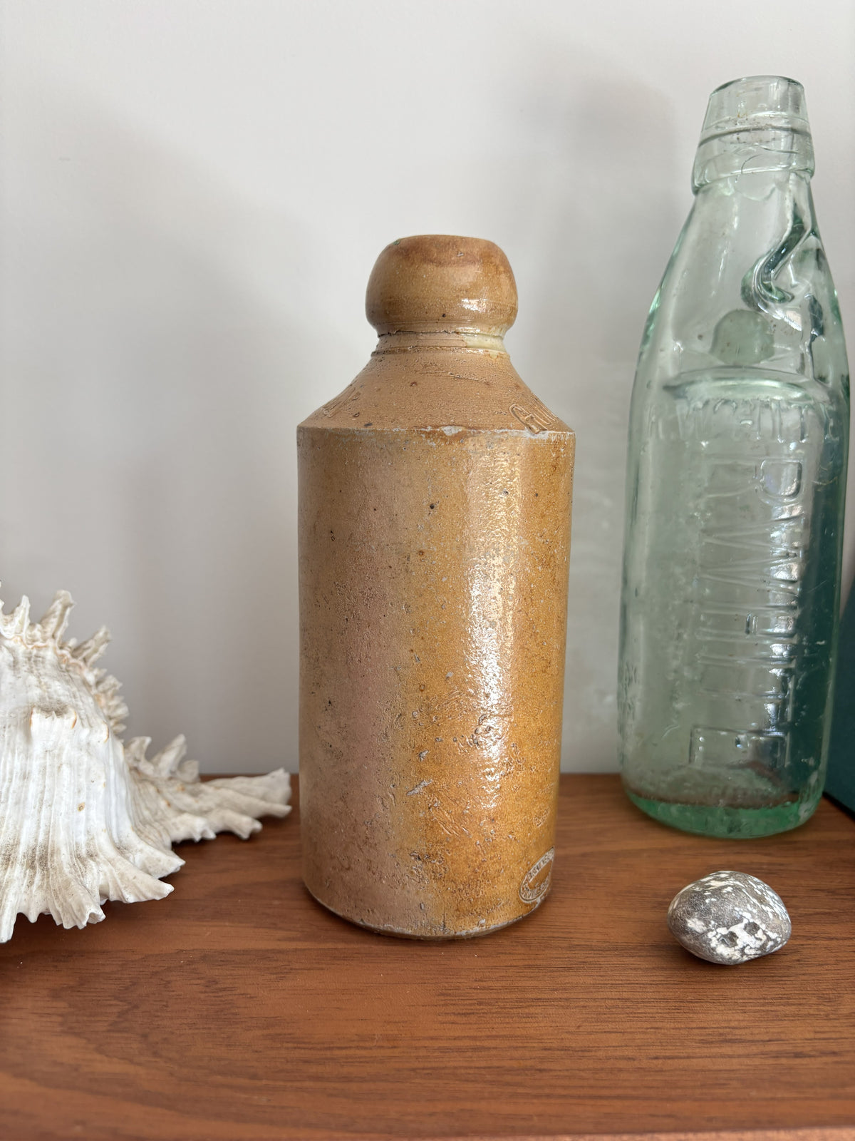 A vintage brown stone bottle with a rounded shoulder and a small top, glazed in a warm sandy tan color, displayed on a wooden surface next to a vintage glass bottle and a seashell.