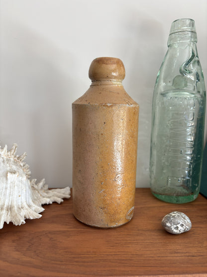 A vintage brown stone bottle with a rounded shoulder and a small top, glazed in a warm sandy tan color, displayed on a wooden surface next to a vintage glass bottle and a seashell.
