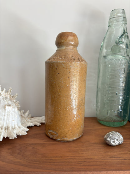 A vintage brown bottle with a rounded top, displayed on a wooden surface next to a vintage green glass bottle, a white seashell, and a small silver rock against a neutral background.