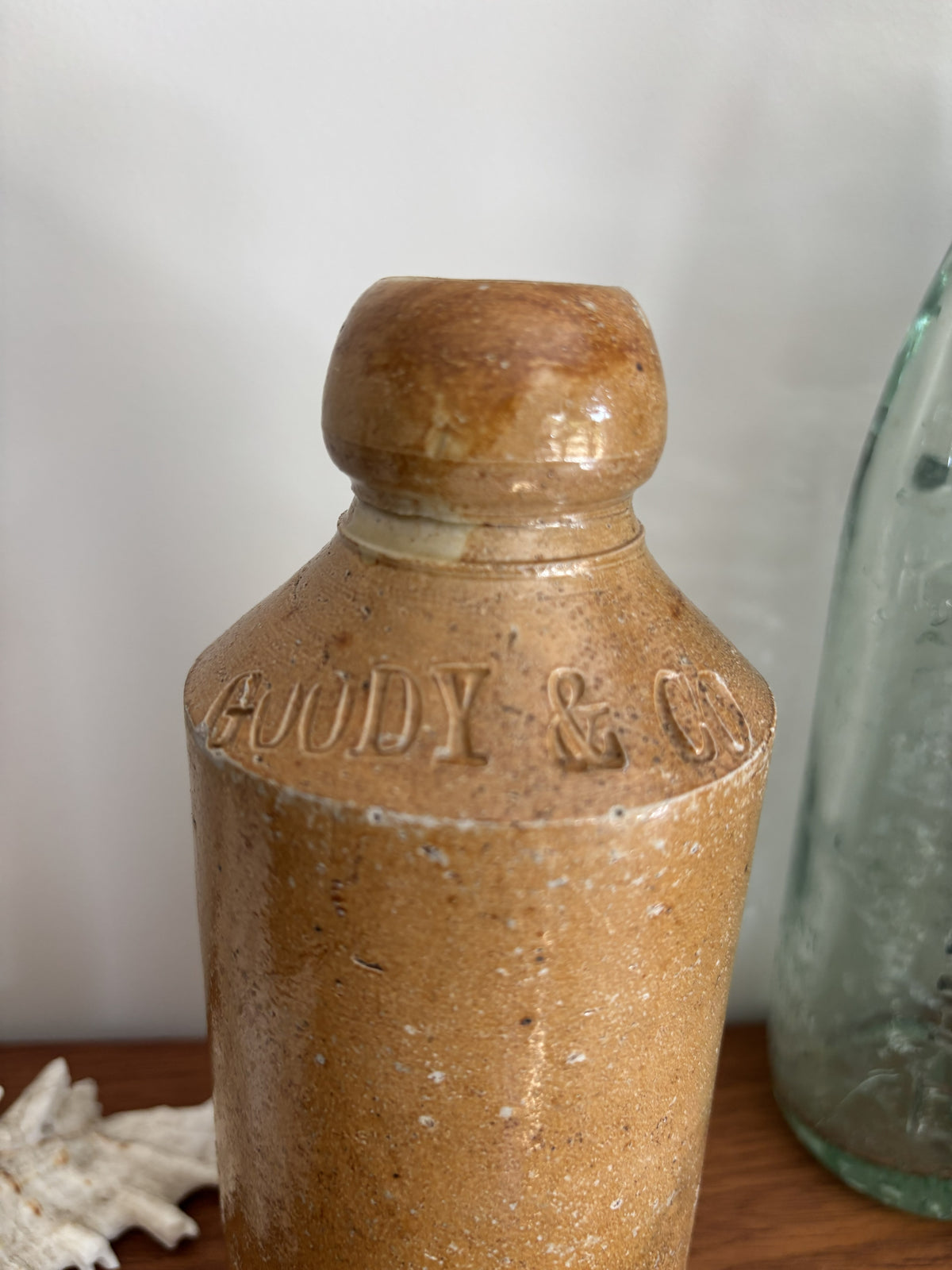 A vintage stoneware bottle with a rounded shoulder embossed with the words Goody & Co on the neck. The bottle has a speckled, earthy brown and tan glaze and is shown on a wooden surface next to a glass shell.