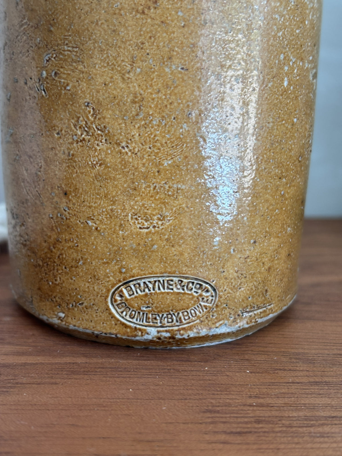 A close-up of a vintage stoneware bottle with a textured brown and gold glaze, featuring an embossed 'Brayne & Co. Stoneware Company' logo near the base, resting on a wooden surface.