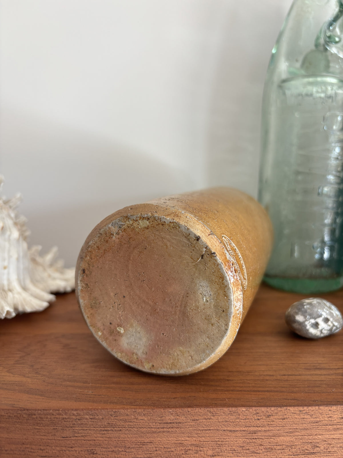 A vintage stoneware bottle base with a textured, earthy brown and beige glaze, shown lying on its end on a wooden surface next to a glass bottle and a shell.