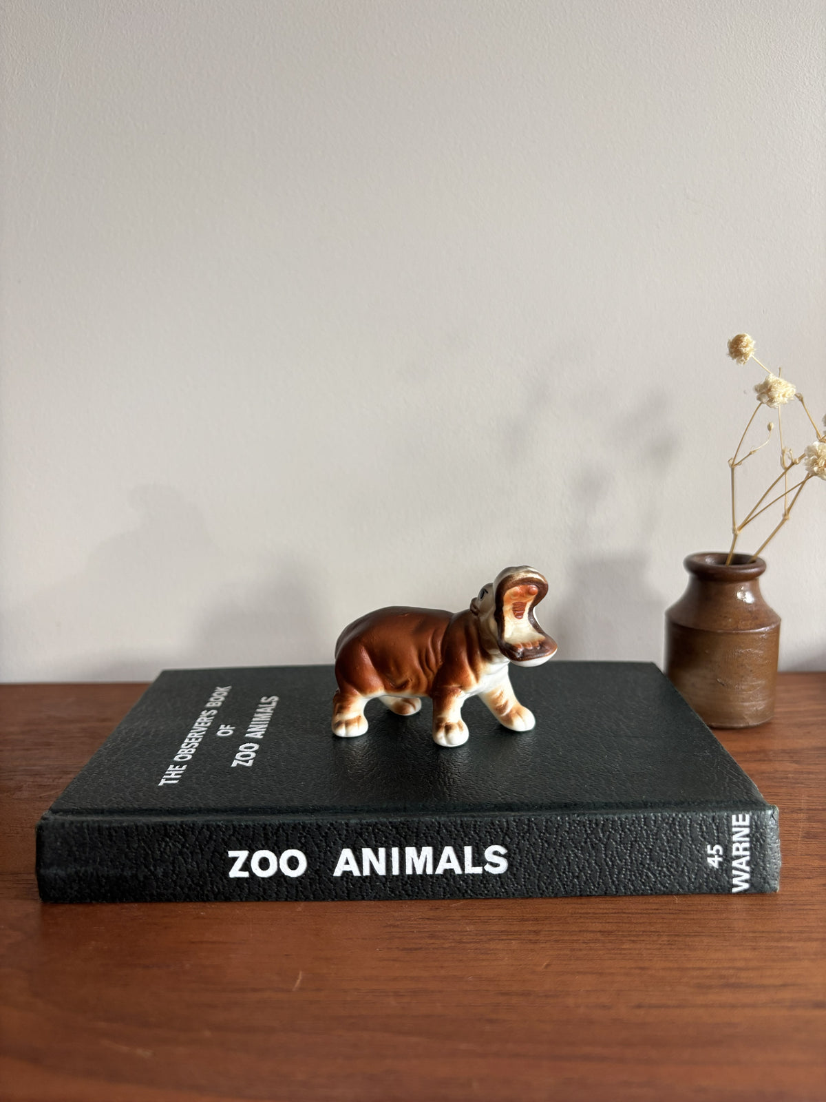 A small ceramic hippopotamus figurine with brown and white coloring stands on the spine of a black book titled 'ZOO ANIMALS' by E. Warne.