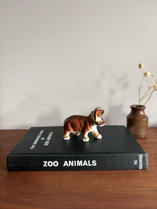 A small ceramic hippopotamus figurine with brown and white coloring stands on the spine of a black book titled 'ZOO ANIMALS' by E. Warne.