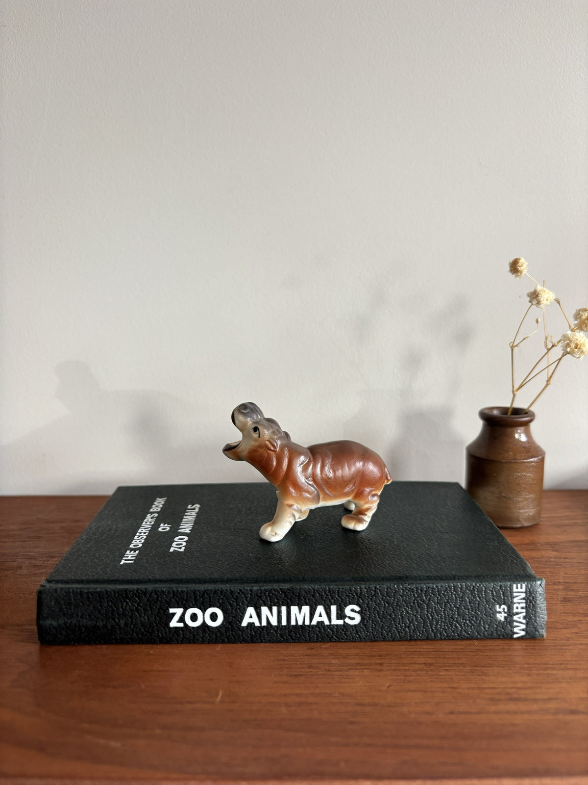 A small brown and white hippopotamus figurine stands on top of a black book titled 'ZOO ANIMALS' on a wooden surface, with a small vase of dried stems against a plain wall in the background.