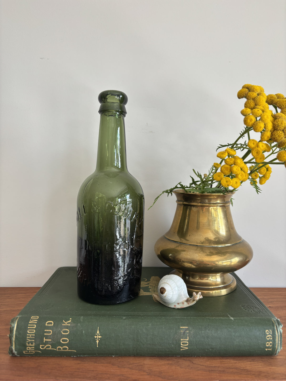 A dark green glass Hole & Compy bottle and a brass-colored vase filled with yellow flowers, styled on top of a vintage green book against a plain light background.