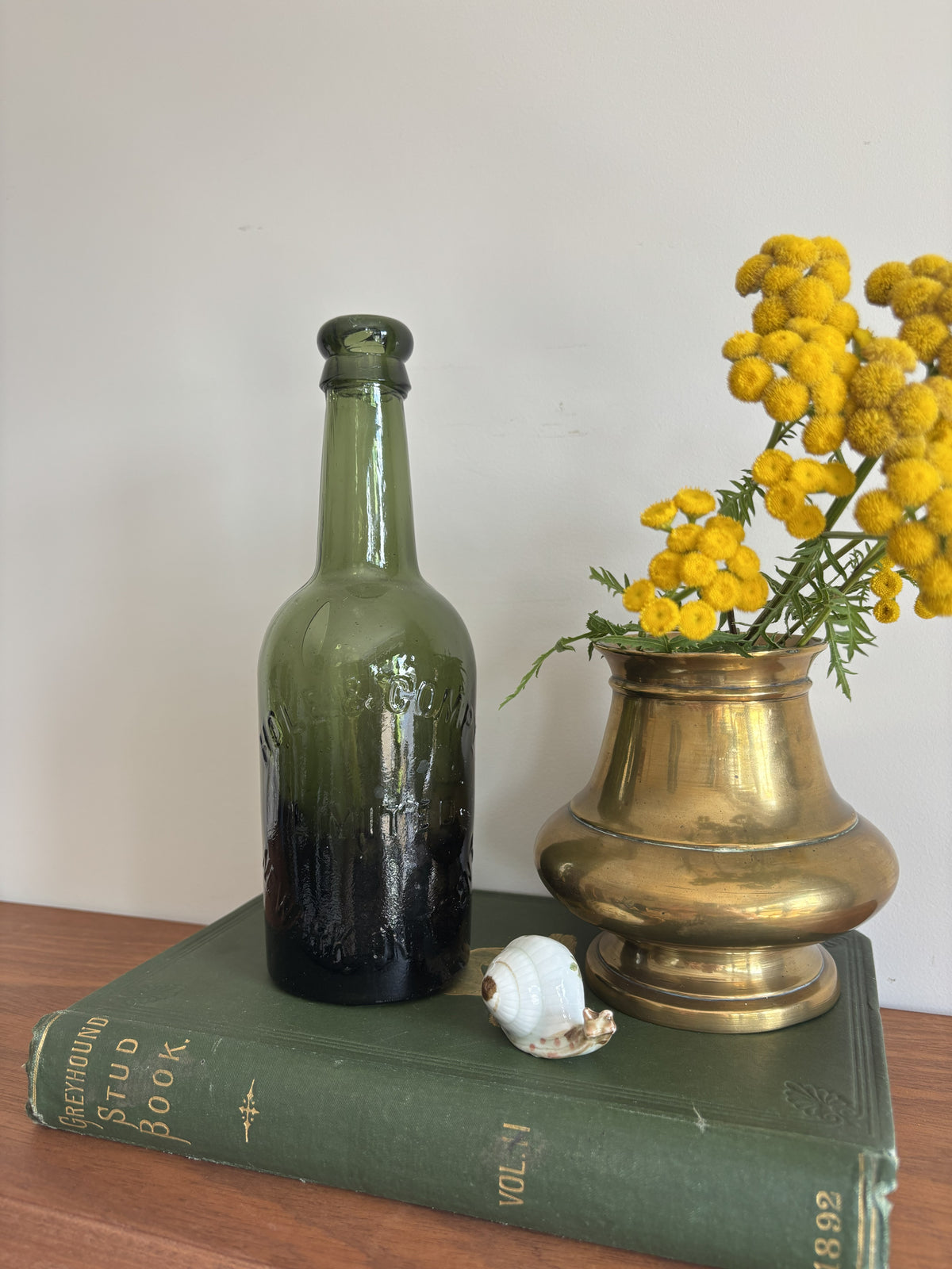 A dark green glass Hole & Compy bottle and a brass-colored vase filled with yellow flowers, styled on top of a vintage green book against a plain light background.