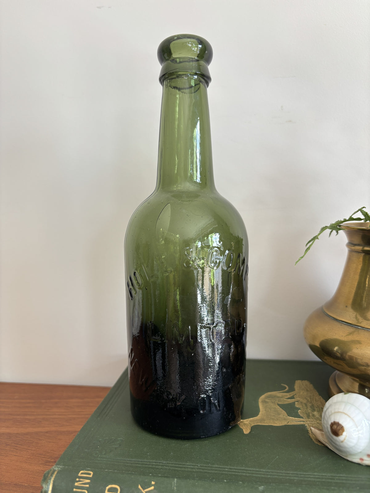 A dark green glass Hole & Compy bottle and a brass-colored vase filled with yellow flowers, styled on top of a vintage green book against a plain light background.