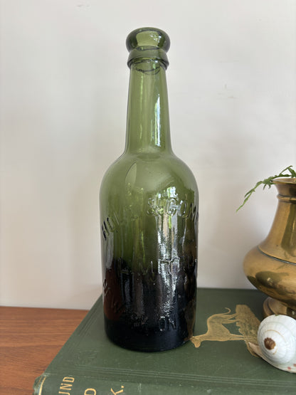 A dark green glass Hole & Compy bottle and a brass-colored vase filled with yellow flowers, styled on top of a vintage green book against a plain light background.