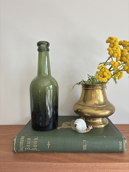 A dark green glass Hole & Compy bottle and a brass-colored vase filled with yellow flowers, styled on top of a vintage green book against a plain light background.
