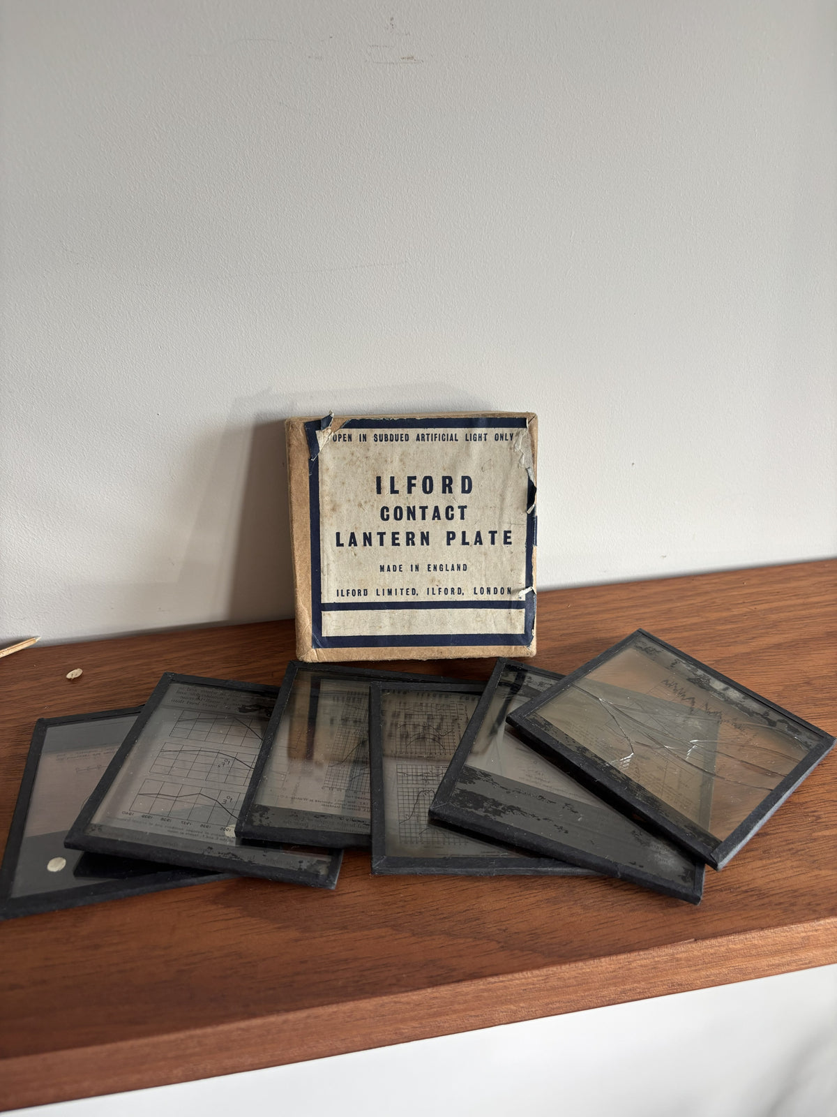 A vintage Ilford Contact Lantern Plate box standing on a wooden shelf, with several square glass contact plates arranged in front of it against a plain light wall.