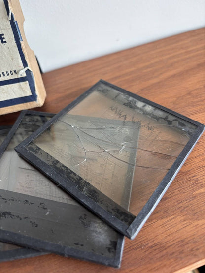 A close-up of vintage lantern plates stacked on a wooden table, with one square piece tilted on top showing aged translucent paper and dark borders.