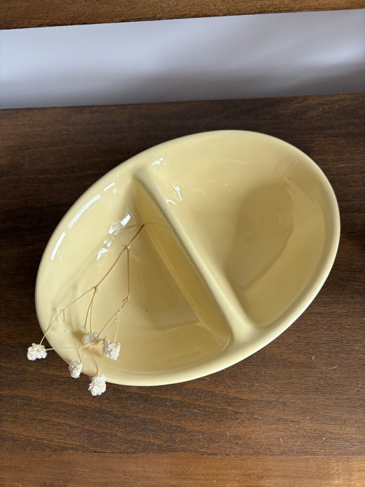 A glossy yellow ceramic divided plate with two sections, shown on a dark wooden surface with small dried white flowers resting in the left side.