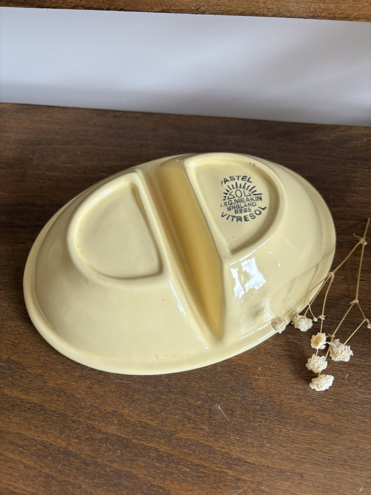 A vintage-style oval divided plate in pale yellow, photographed on a dark wooden surface with a white wall in the background and small dried flowers beside it.