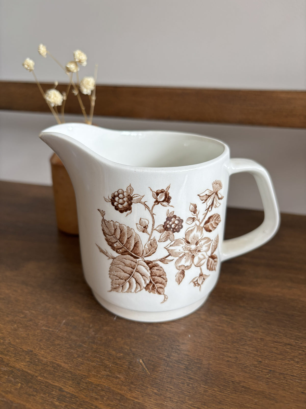 A white ceramic pitcher with a brown floral and berry pattern, set on a dark wooden table with a blurred wooden chair and dried flowers in the background.