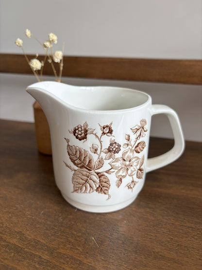 A white ceramic pitcher with a brown floral and berry pattern, set on a dark wooden table with a blurred wooden chair and dried flowers in the background.
