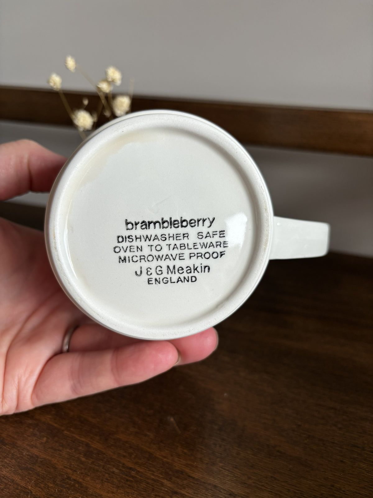 A hand holds a white ceramic mug upside down to show the printed bottom text 'brambleberry J & G Meakin ENGLAND' on a dark wooden surface.