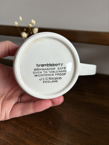 A hand holds a white ceramic mug upside down to show the printed bottom text 'brambleberry J & G Meakin ENGLAND' on a dark wooden surface.