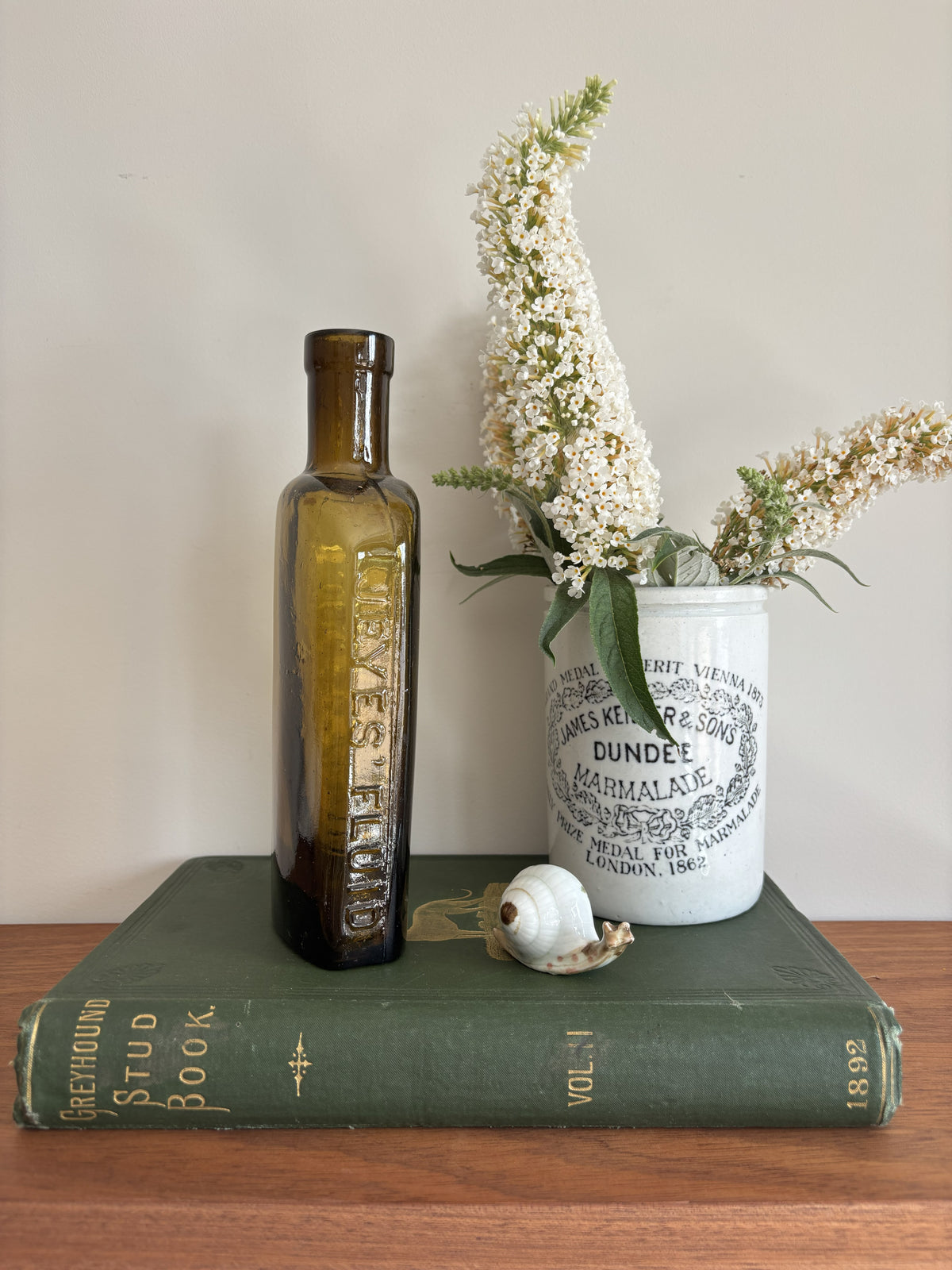 A styled tabletop scene with a tall amber Jeyes' Fluid glass bottle standing on a dark green book, next to a white ceramic jar labeled Dundee Marmalade with white flowers in it