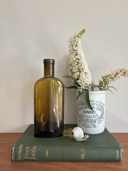 A styled tabletop scene with a tall amber Jeyes' Fluid glass bottle standing on a dark green book, next to a white ceramic jar labeled Dundee Marmalade with white flowers in it