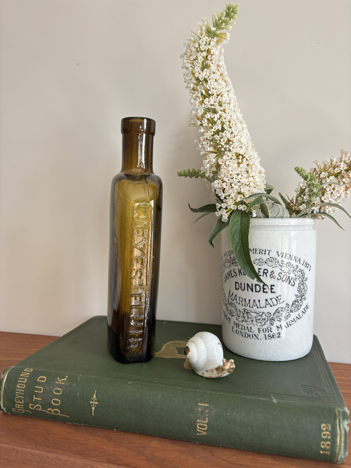 A styled tabletop scene with a tall amber Jeyes' Fluid glass bottle standing on a dark green book, next to a white ceramic jar labeled Dundee Marmalade with white flowers in it