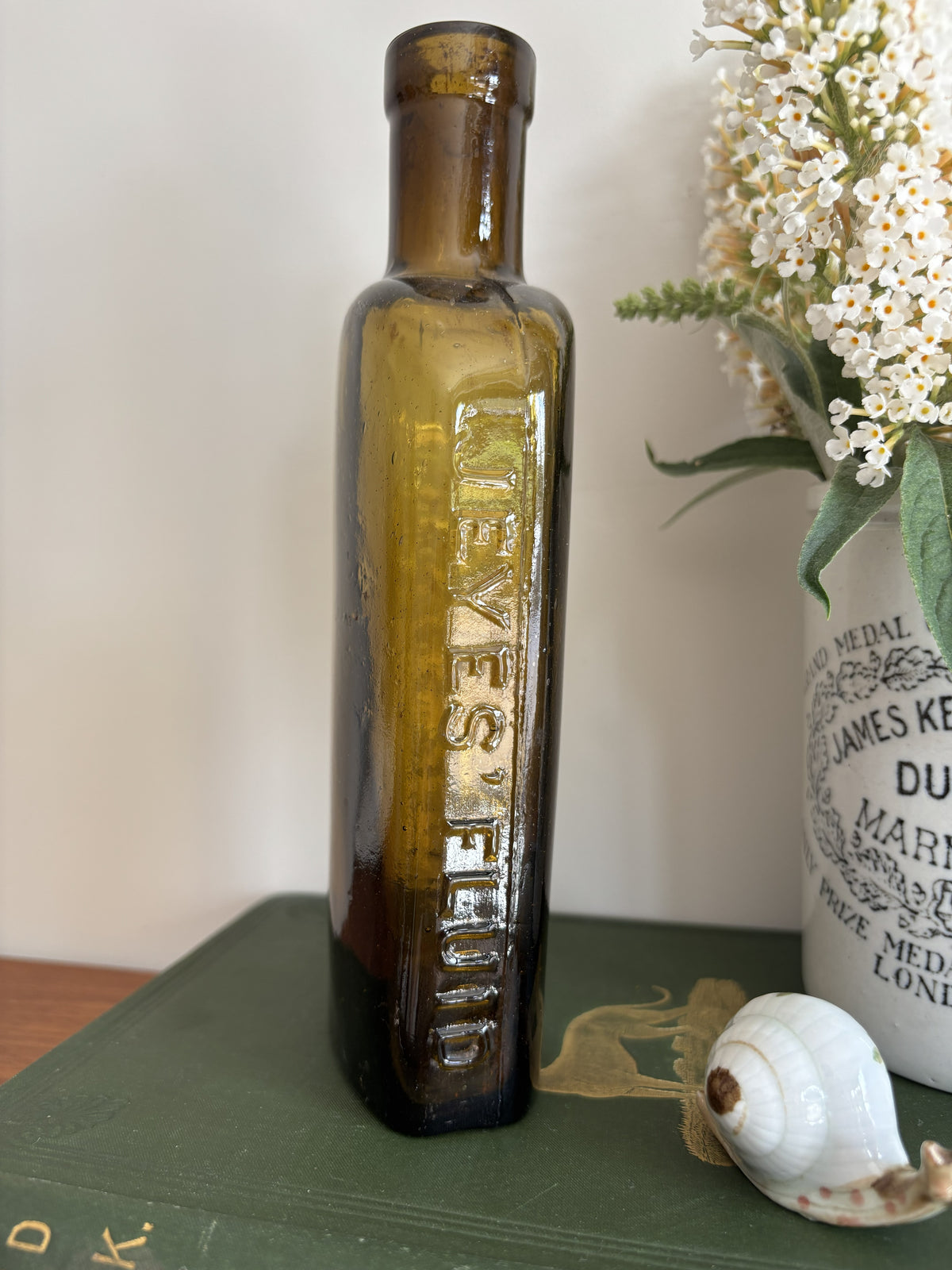 A styled tabletop scene with a tall amber Jeyes' Fluid glass bottle standing on a dark green book, next to a white ceramic jar labeled Dundee Marmalade with white flowers in it