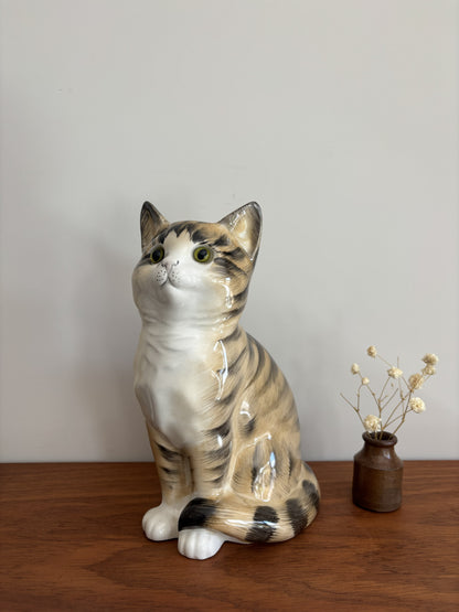 A glossy ceramic cat figurine with tabby and white coloring, sitting on a wooden surface against a plain light gray wall, with a small vase of dried stems beside it.