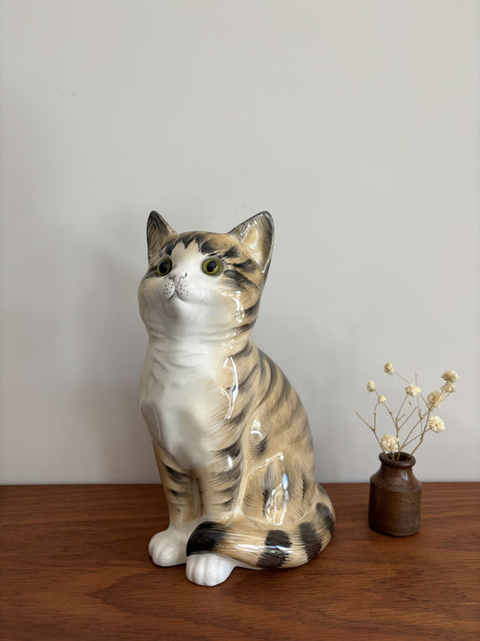 A glossy ceramic cat figurine with tabby and white coloring, sitting on a wooden surface against a plain light gray wall, with a small vase of dried stems beside it.