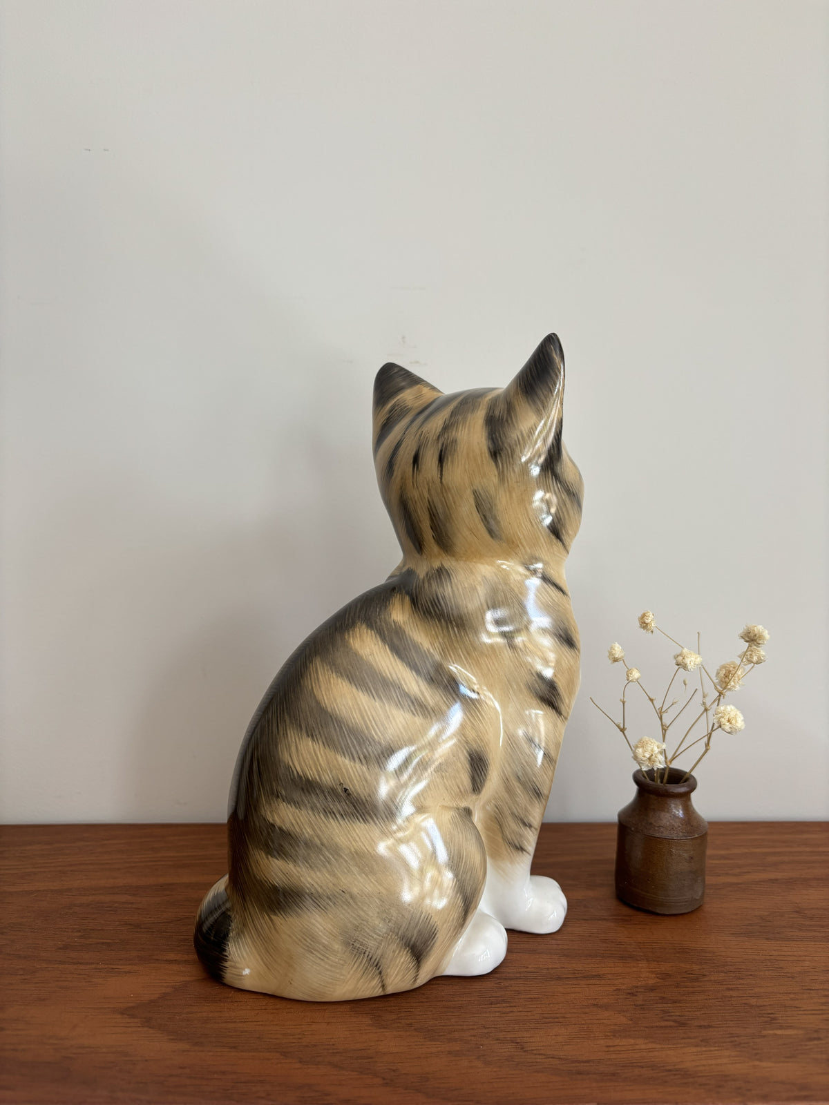 A glossy ceramic cat figurine in a seated position, shown from the back on a wooden surface against a plain light gray wall, with a small brown vase of dried stems beside it.
