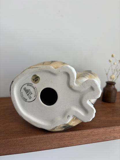 A vintage ceramic bird figurine shown from its underside, resting on a wooden shelf against a plain light gray wall. The base features a white glazed finish with a circular black stamp reading 'Just Cats & Co Staffordshire England'.