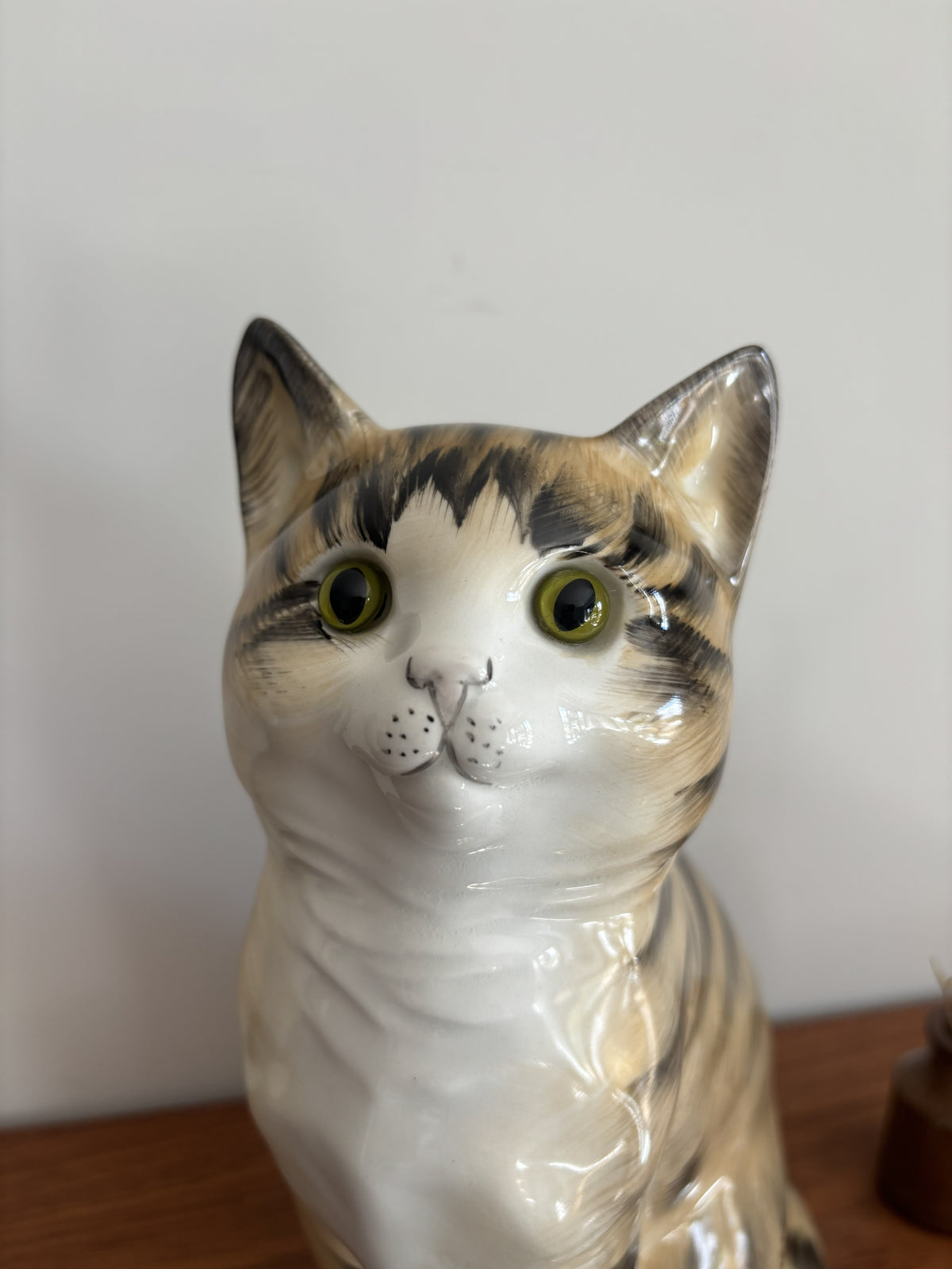 A glossy ceramic cat figurine with a white, gray, and black tabby pattern, featuring large yellow-green eyes, shown on a wooden surface against a plain light background.
