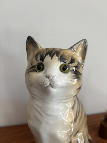 A glossy ceramic cat figurine with a white, gray, and black tabby pattern, featuring large yellow-green eyes, shown on a wooden surface against a plain light background.