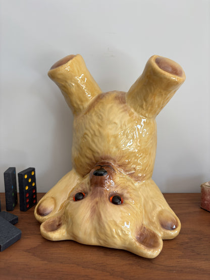 A glossy ceramic teddy bear in an upside down pose, displayed on a wooden surface against a plain light wall.