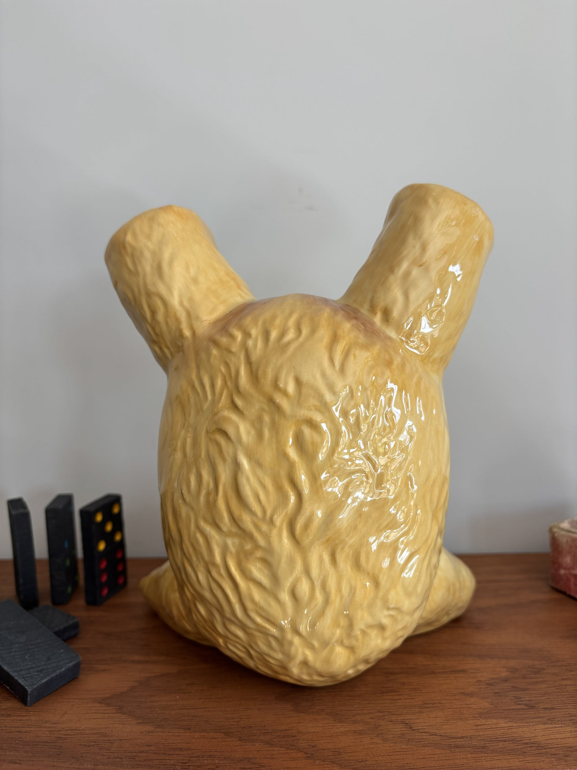 The reverse of a glossy ceramic teddy bear in an upside down pose, displayed on a wooden surface against a plain light wall.