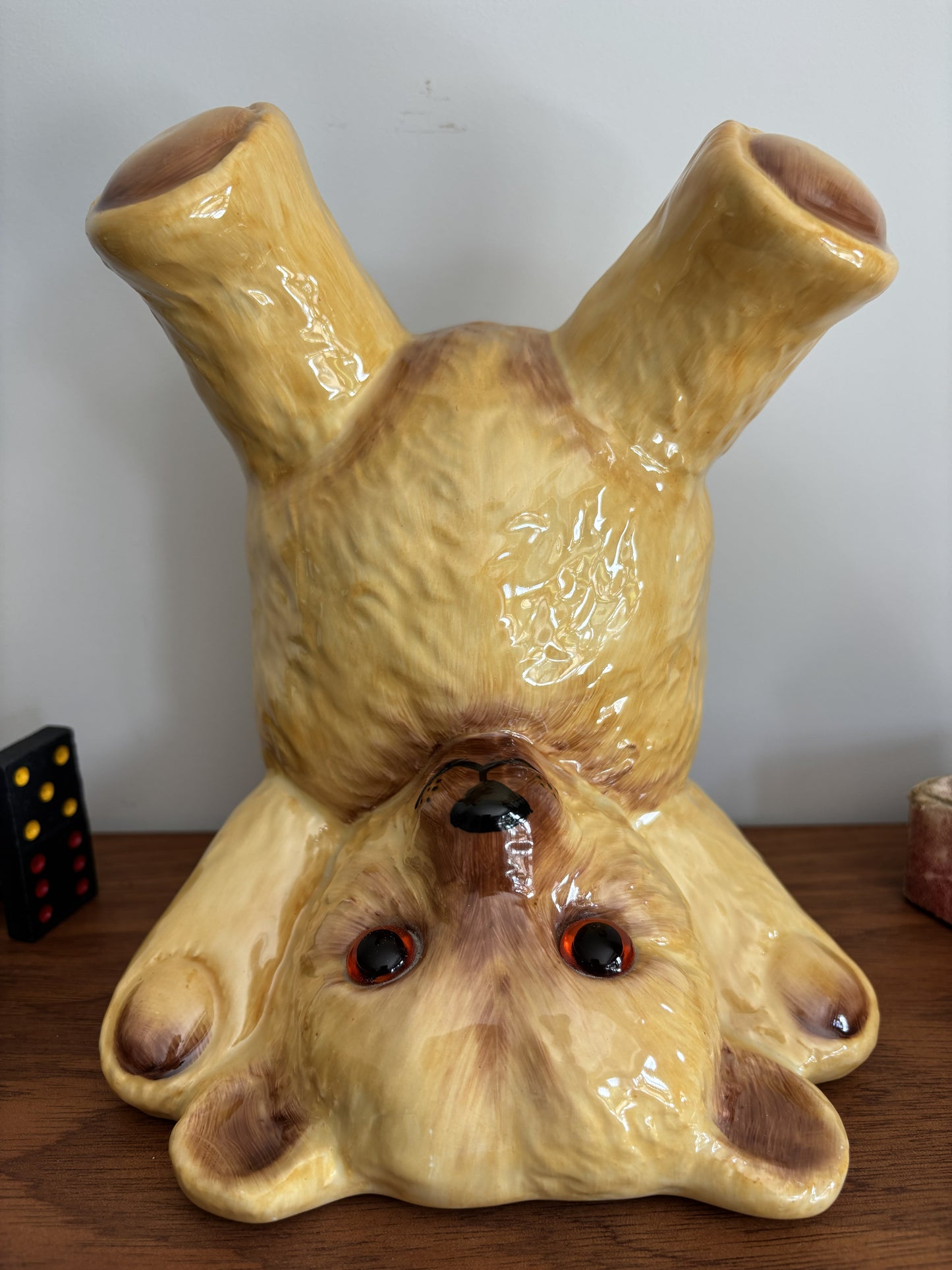 A glossy ceramic teddy bear in an upside down pose, displayed on a wooden surface against a plain light wall.