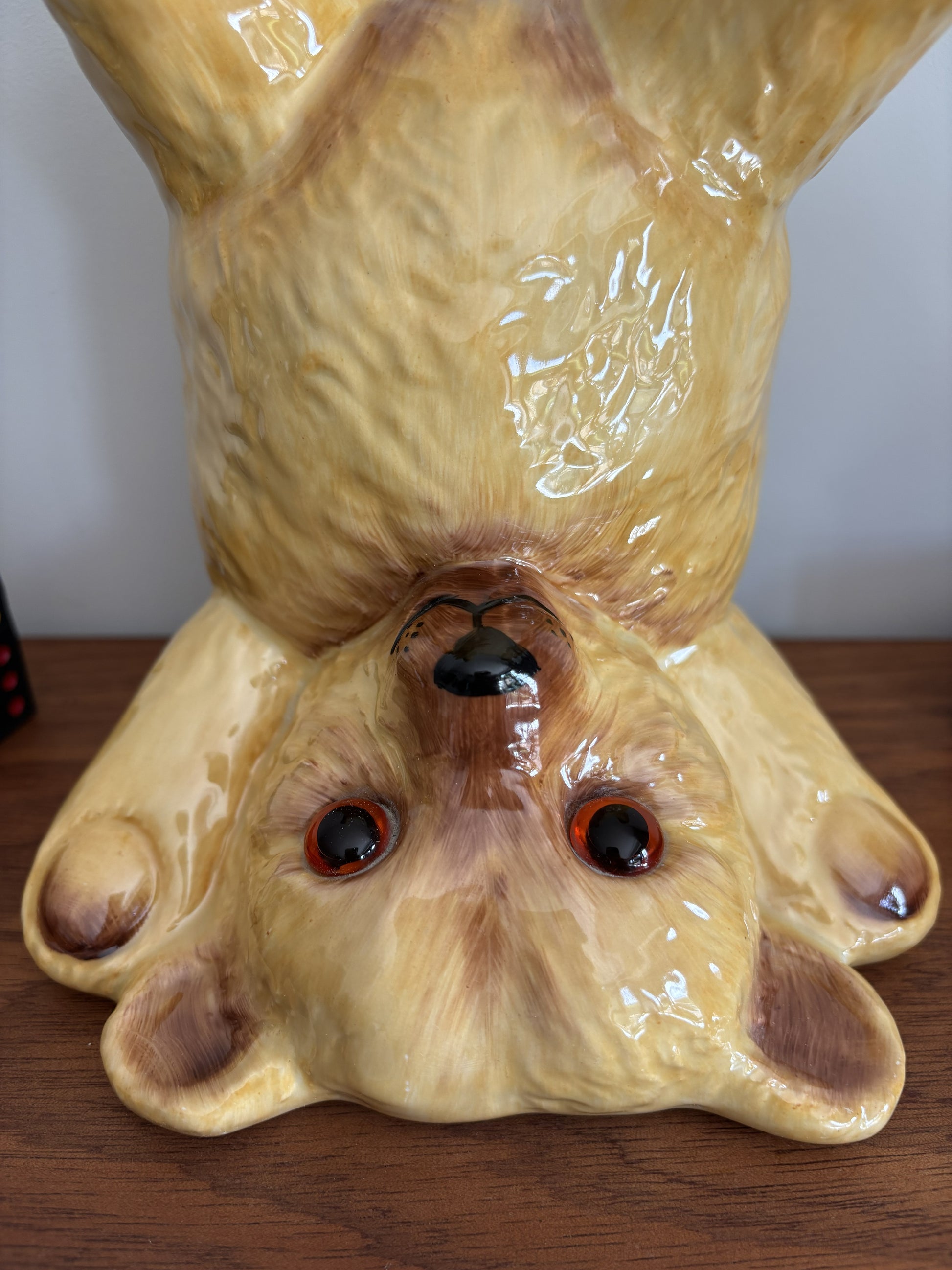 A facial close up of glossy ceramic teddy bear in an upside down pose, displayed on a wooden surface against a plain light wall. 