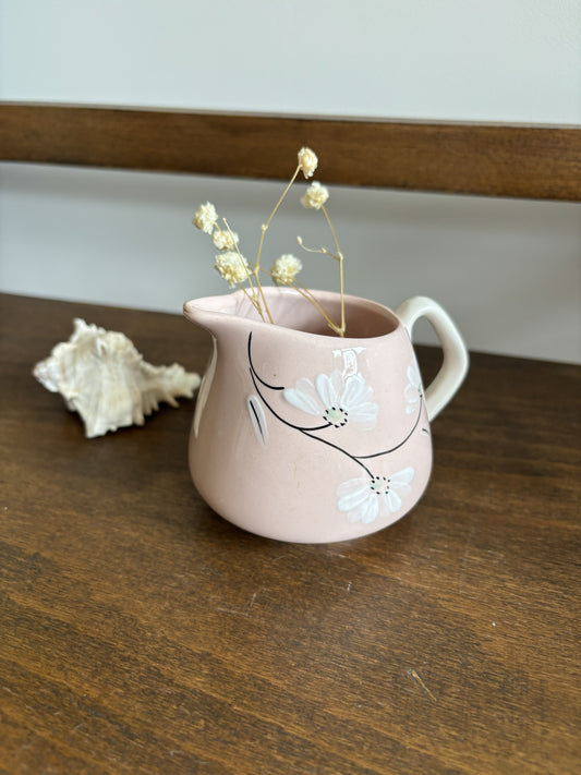 A small dusty pink ceramic pitcher with white floral decals and dried baby's breath stems, set on a dark wooden table next to a white seashell.