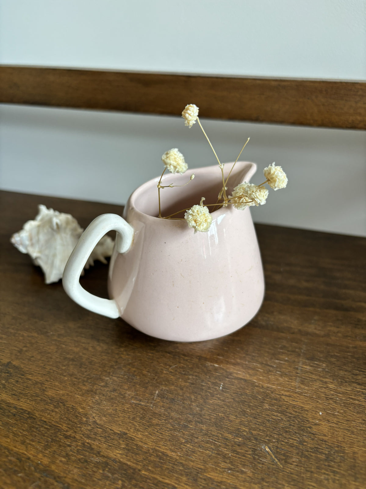 A small speckled ceramic pitcher in a dusty pink color with a white handle, sitting on a dark wooden table. It is styled with dried baby's breath flowers and a white seashell.
