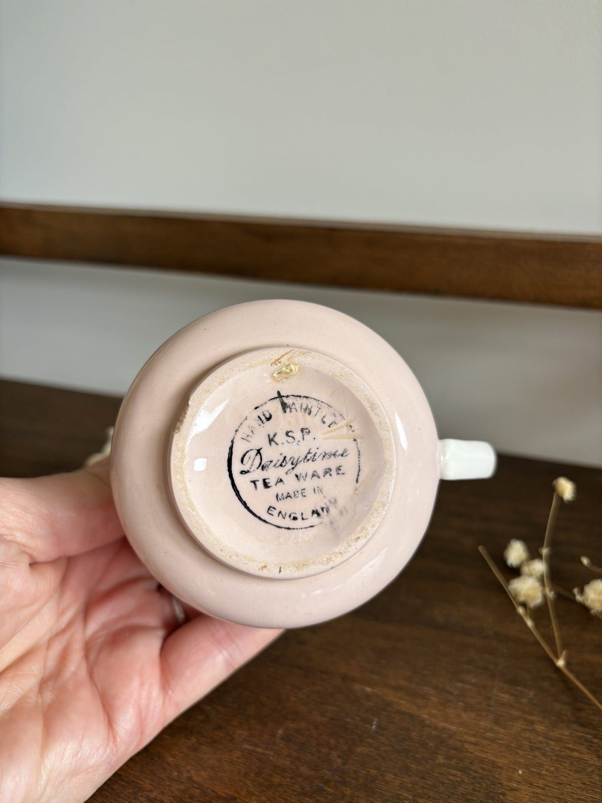 A hand holding the base of a small pale pink ceramic teacup or saucer-shaped tea holder, showing a printed circular maker's mark that reads 'K.S.P Daisytime Tea Ware'