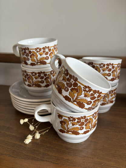 A set of vintage ceramic mugs and saucers with a white base and a brown floral pattern, arranged on a wooden table with dried flowers in the foreground.