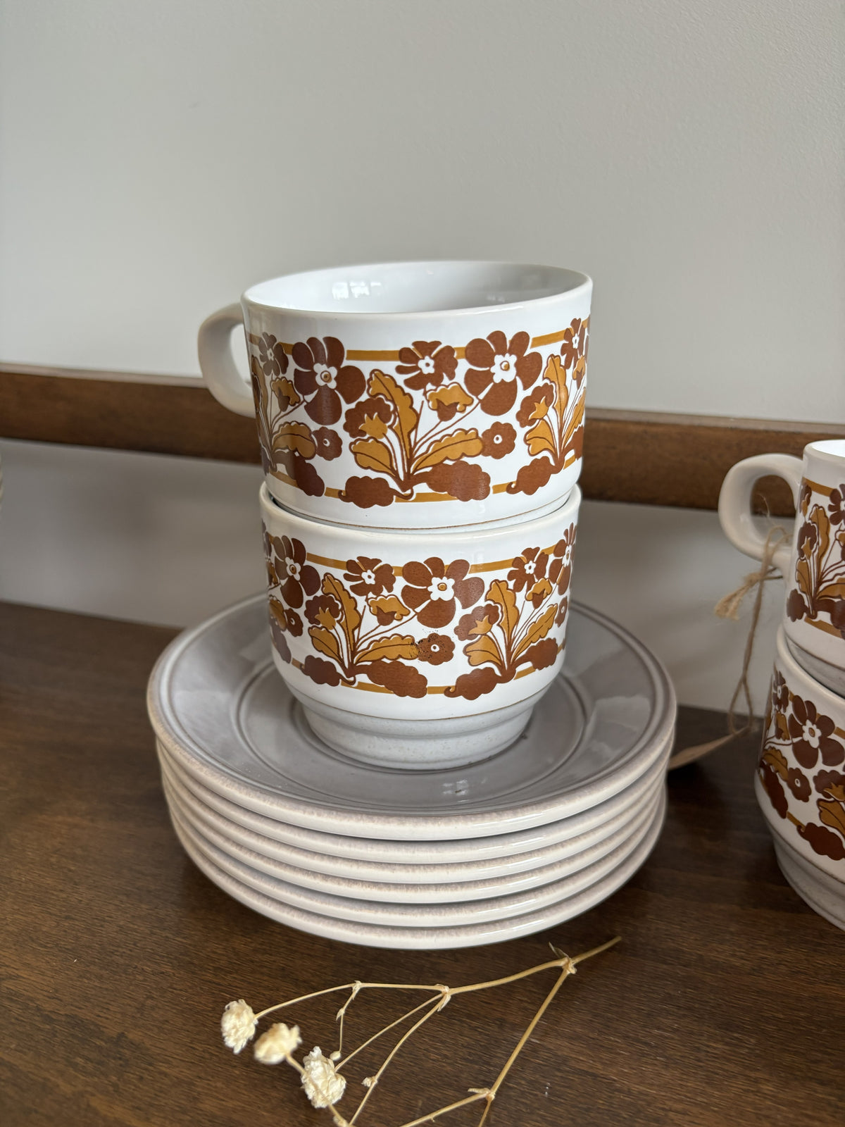 A stack of white ceramic cups and saucers with a brown and gold floral pattern, displayed on a wooden surface.