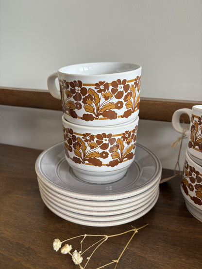 A stack of white ceramic cups and saucers with a brown and gold floral pattern, displayed on a wooden surface.