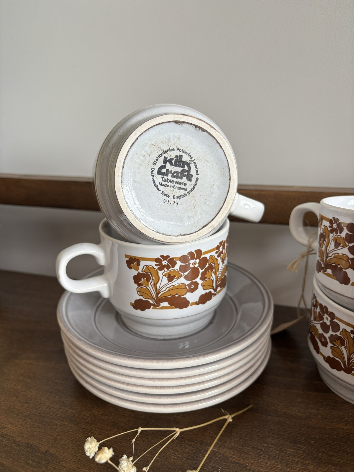 A stack of white ceramic teacups with saucers and handles, decorated with a brown botanical leaf pattern, displayed on a dark wooden surface against a neutral background.