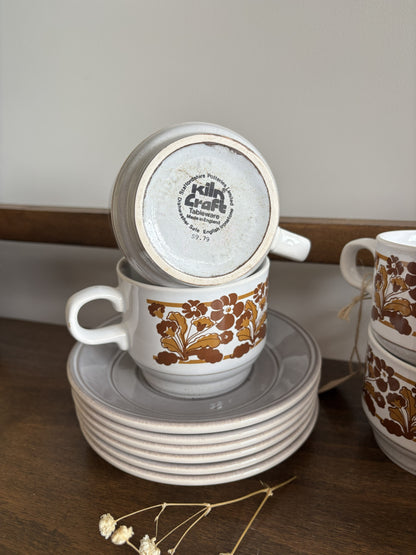 A stack of white ceramic teacups with saucers and handles, decorated with a brown botanical leaf pattern, displayed on a dark wooden surface against a neutral background.